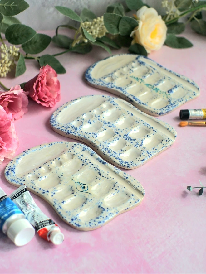 a trio of ceramic paint palettes, handcarved from stoneware clay and glazed in a splatter glaze with blue and white speckles. Each paint palette has a different design of bird, goddess or plain. Photographed with pink flowers on a pink background, surrounded by paints, Jenny Hopps Pottery, Ceramic gifts, artist gifts, painter gifts, creative gifts