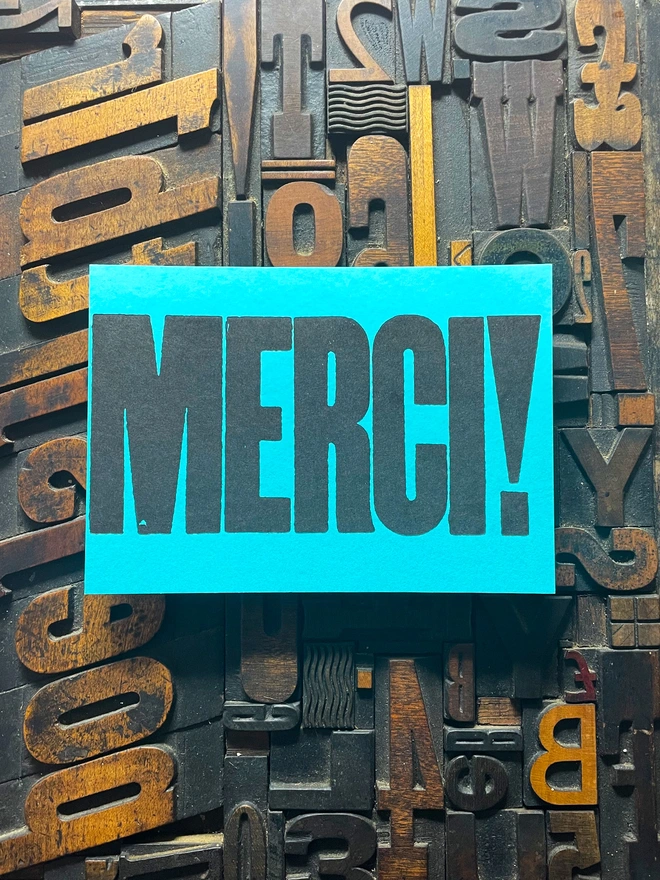 A bright turquoise thick duplex card with the word "MERCI!" printed in bold, black letters lies on a surface covered with various letterpress blocks.