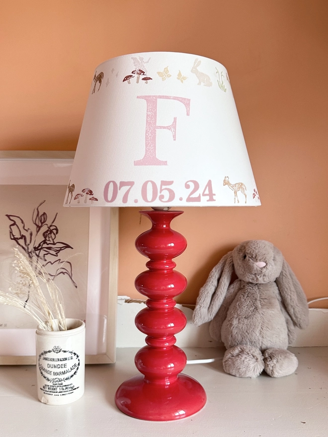 Personalised kids lampshade with woodland animals design and pink initial and date, styled on a red lamp base in a girl’s bedroom. A pretty empire lampshade ideal for kids bedroom décor and new parent gifts.