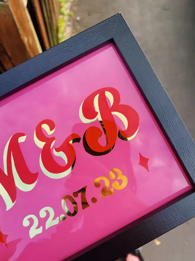 Close-up of the black framed 'M & B' wedding sign with a curly script style on pink background, with red lettering and gold leaf shade and date.