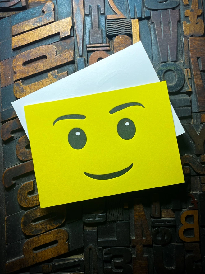 A bright yellow letterpress card with a smiling face emoji. The background consists of various wooden printing letters in different orientations and sizes.