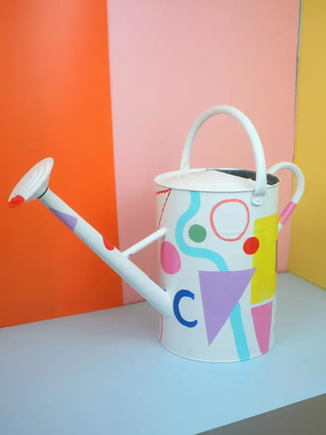 Hand painted 12 litre watering can by Julie-Anne Pugh. Base colour is white with bring coloured shapes weaving across the body of the can on a yellow and orange background. 