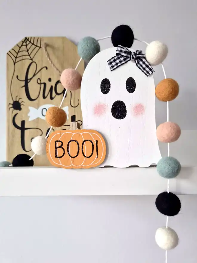 A wooden Halloween ghost on a shelf, with a felt ball garland draped over it in muted tones.