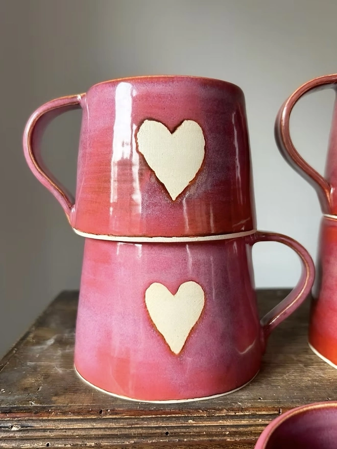 Heart Mug Raspberry Red, a pair of red heart mugs on a wooden surface stacked on top of each other. 