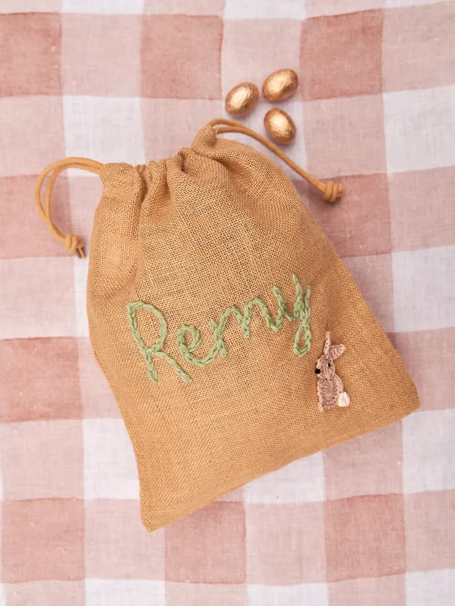 jute drawstring sack with brown knitted bunny decoration and green 'remy' name next to golden chocolate eggs on red gingham table cloth