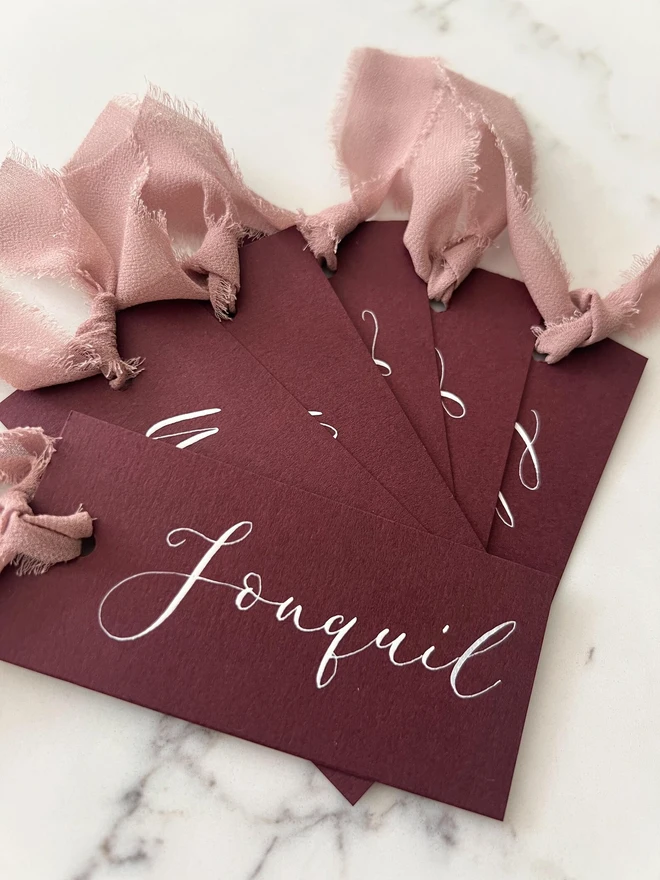 Personalised Calligraphy Wedding Burgundy Place Cards, burgundy place cards set together on a wooden table with chiffon plum ribbons. 