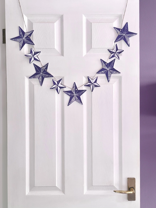 'Bright Star' Colourful Hand Printed Linocut Garland Hanging In Front Of A White Door Purple Star Mix Garland Of Large And Small Stars