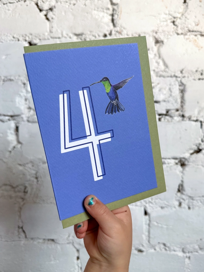 a deep purpley blue card with a big white number 4 and a hummingbird illustration and a green envelope