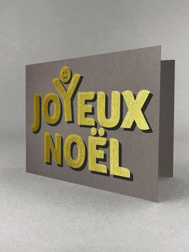Golden text screenprinted onto grey card stock, spells out Joyeux Noel with a happy face above the Y, as though celebrating. Landscape card stood on a grey background, slightly angled.