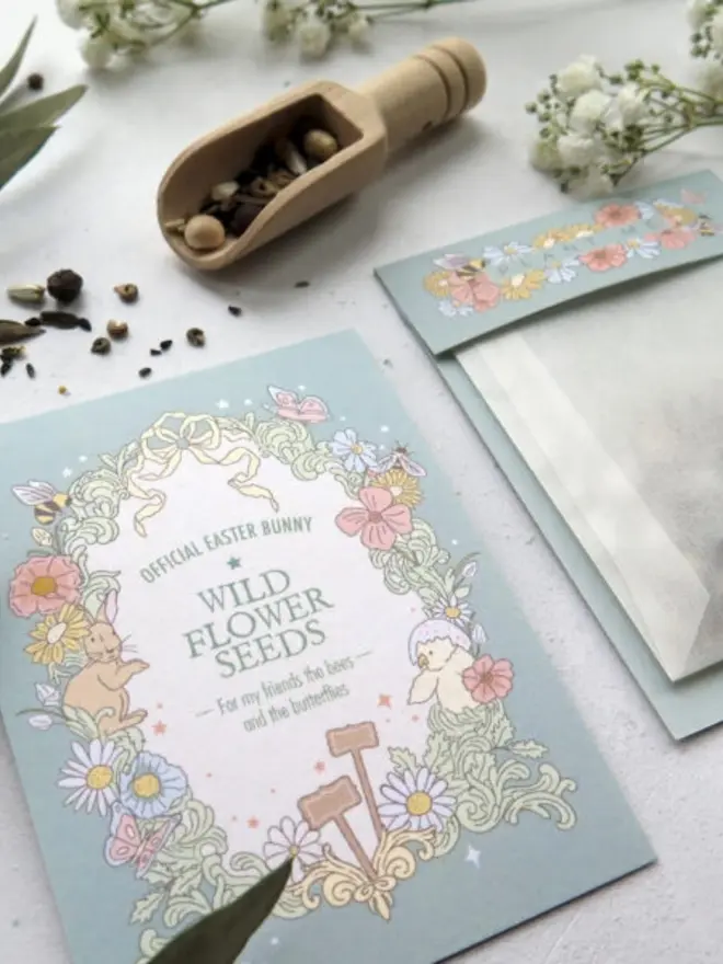 Close-up of the wildflower seed card, with a pastel border of poppies, daisies, a bunny and a chick. A wooden scoop of mixed seeds and the "Plant Me" pouch are behind it.