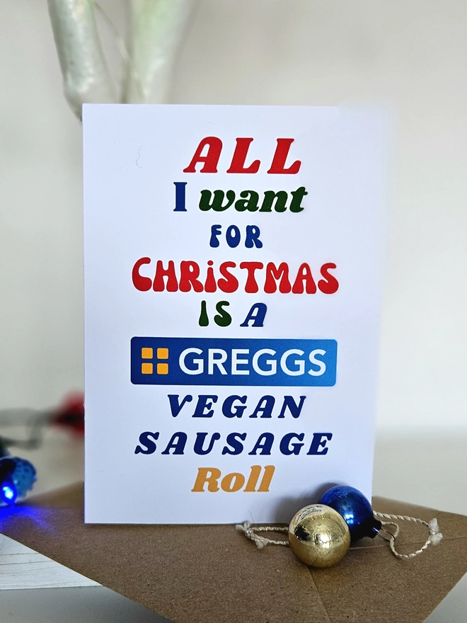 A colourful card Christmas Card that says All I want for Christmas is a Vegan Sausage Roll in retro style fonts on a white background.