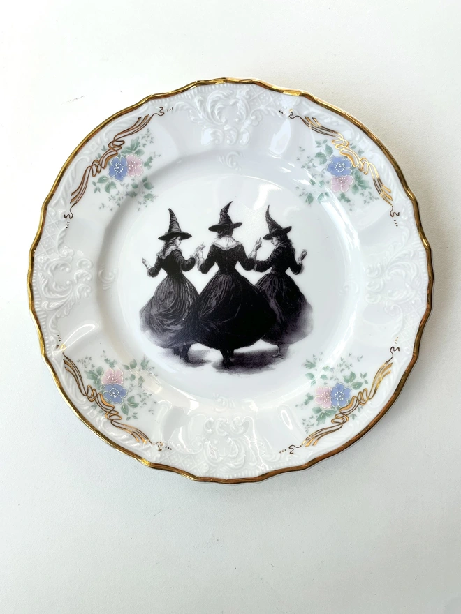 a coven of 3 witches dance hand in hand, the image is victorian in black and white and is printed directly onto an ornate vintage plate