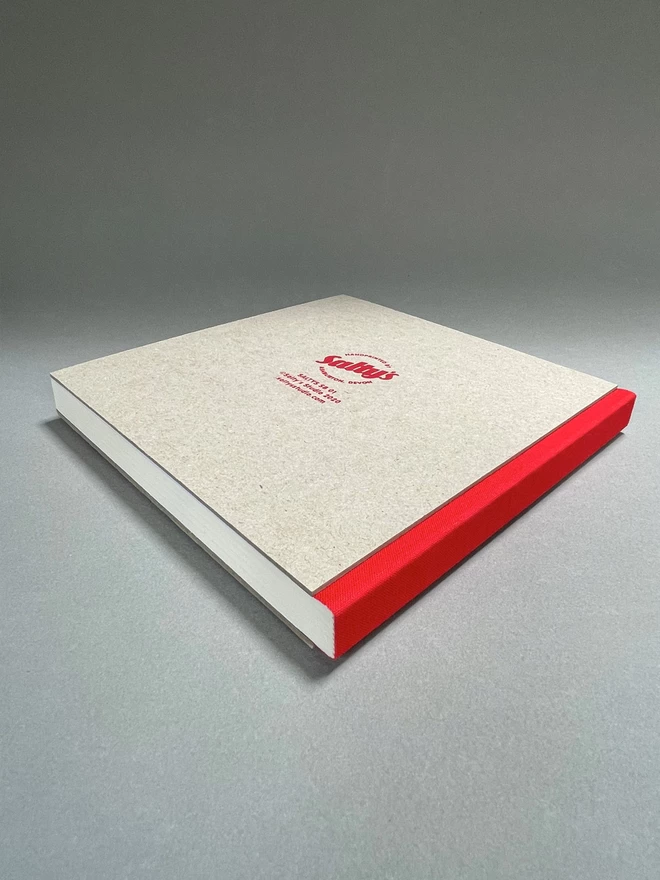 Rear view of a pasteboard sketchbook with a red spine and a saltys studio brand logo on reverse, lies on a light grey studio backdrop. Red fabric spine towards us.