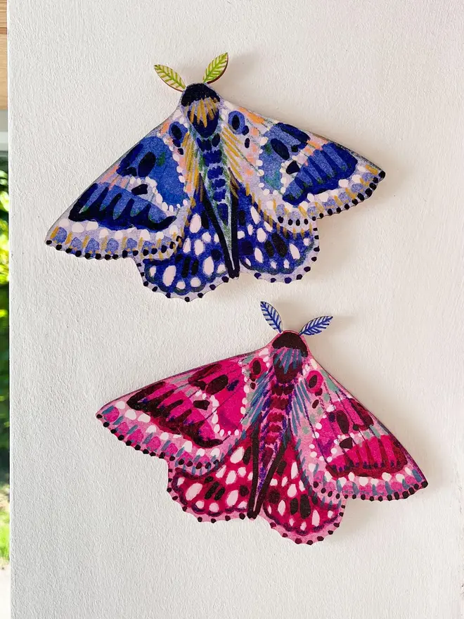wooden moth wall decoration. pink and blue wooden moth decorations hung on plain white wall.