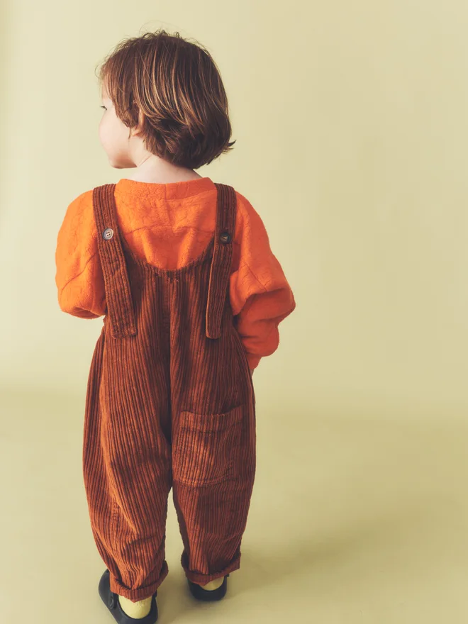 Back view of a toddler against a pale yellow backdrop, showing the button straps and rear patch pocket of Studio Koter rust corduroy Kai dungarees, worn over the Riverson top in Teddy Orange