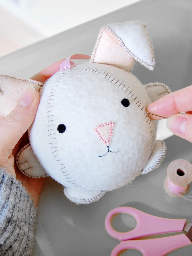 A white felt rabbit toy is being sewn together, with pink scissors and various craft kit components on the grey surface below.