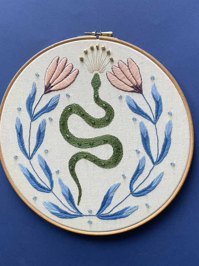 Embroidery hoop containing a green snake blue leaves pink flowers gold crown and beads