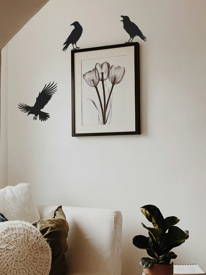 Black picture frame above a cream sofa, the picture frame has two black crows on the top of each corner and at the left side is a black flying crow
