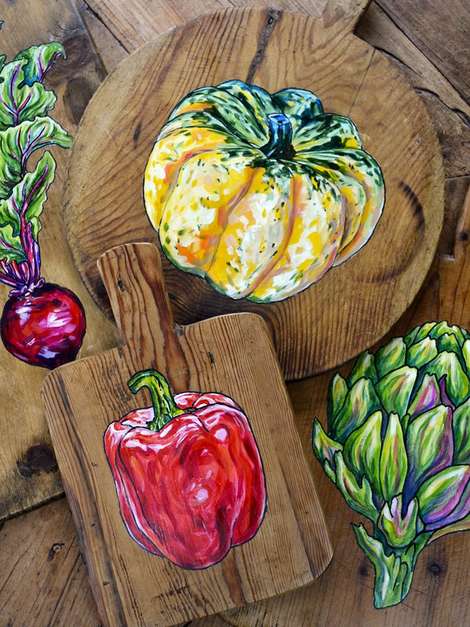 A collection of wooden chopping boards with different handpainted designs including a squash, a red pepper, an artichoke and a beetroot