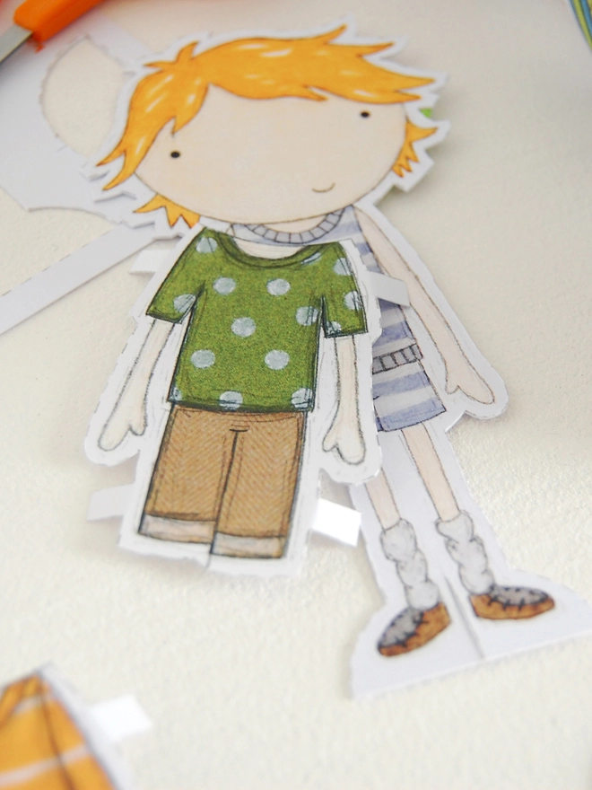A paper doll with cut out outfits, from wrapping paper, lays on a white table.