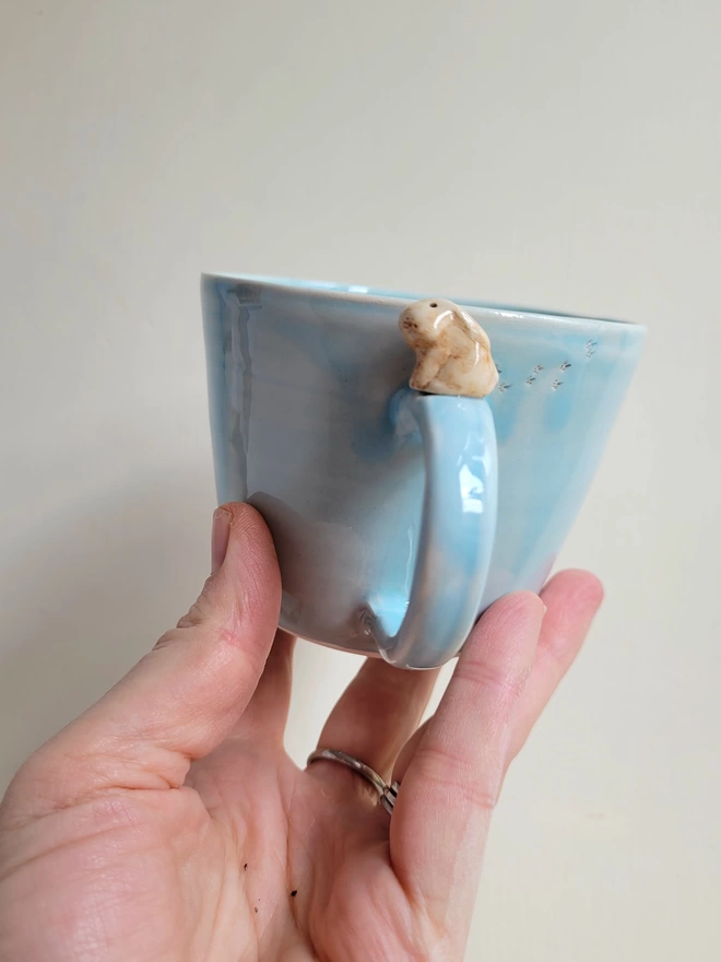 Light turquoise blue ceramic cup held in a hand there is a sweet bunny rabbit on the handle and footprints on the cup