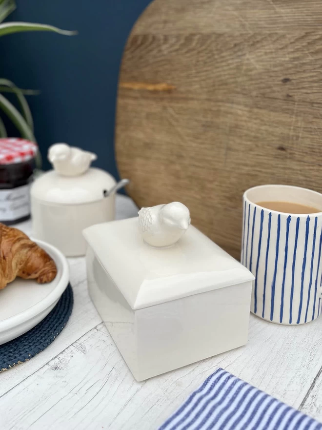 A handmade ceramic butter dish sits with a sugar pot both with matching bird handles.