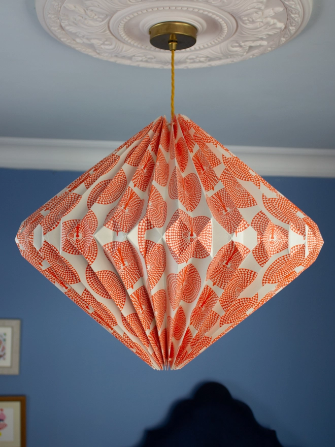 Handmade origami diamond shaped paper lightshade featuring a hand illustrated orange African sun mask design.