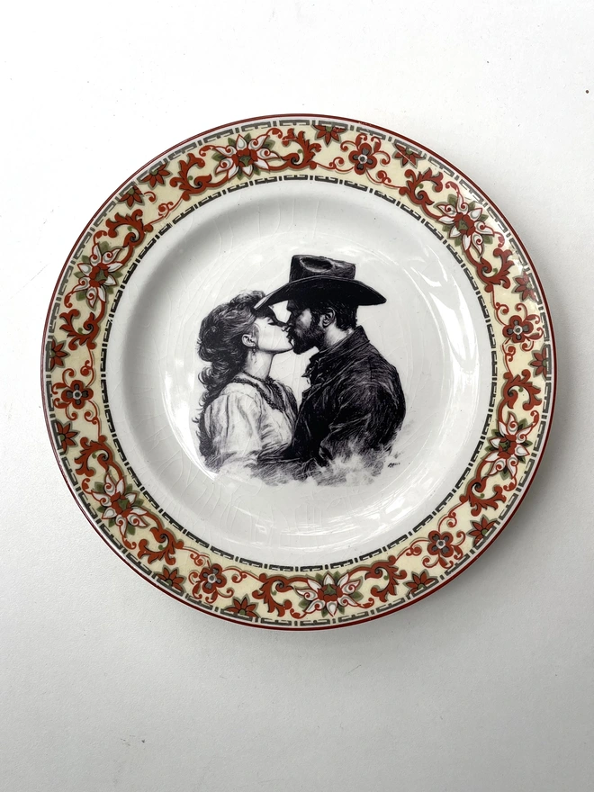 a vintage plate with an ornate border has a black and white vintage illustration of a cowboy and cowgirl sharing a kiss in the centre