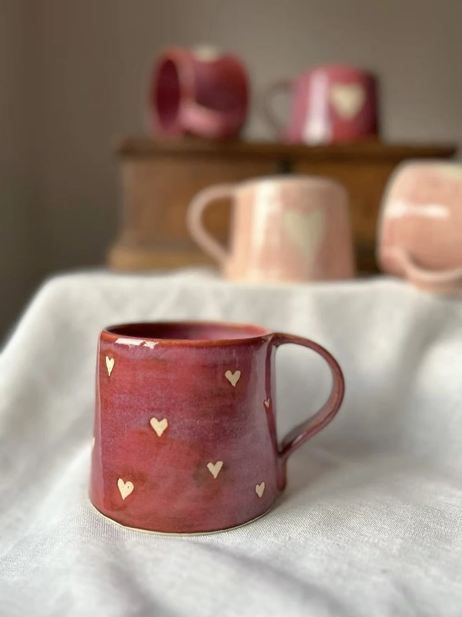 Love Heart Mug Raspberry, red heart mug on a white surface in front of other mugs in the background on a wooden surface. 