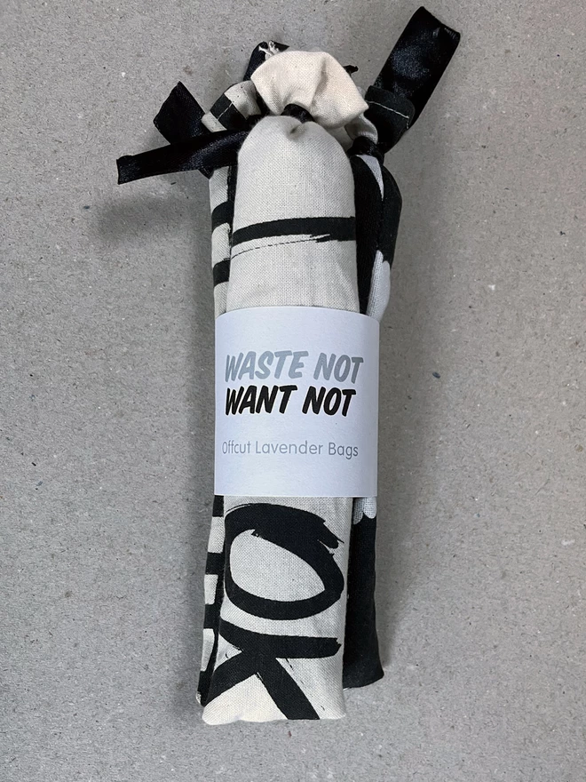 A bundle of three long style cotton lavender bags, tied at the tops, gathered with a branded cuff, sit on a greyboard background - it says Waste Not Want Not, Offcut Lavender Bags. The fabric has bits of black and white printed pattern visible.