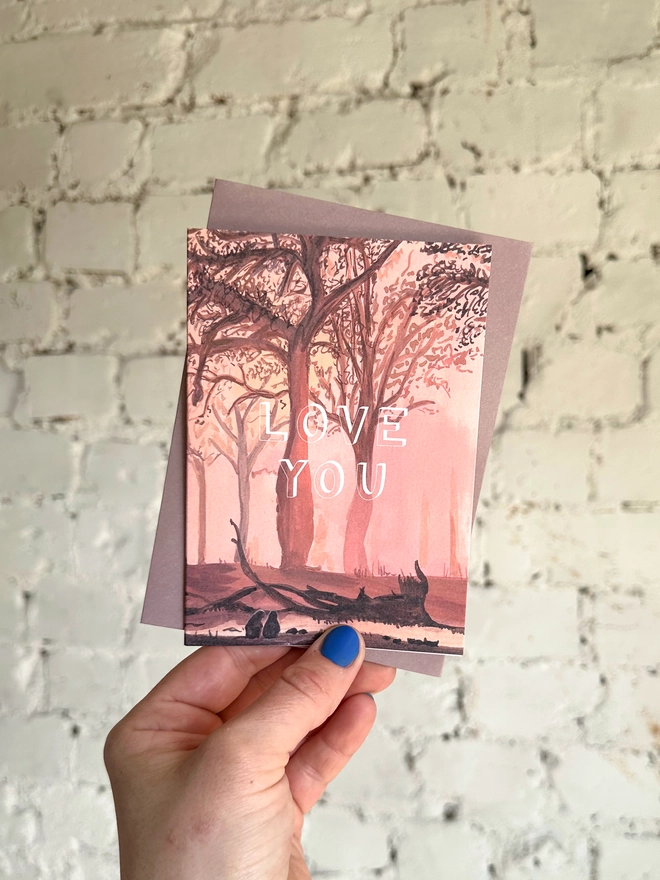 a greetings card featuring an illustration of a sunset through the trees with the text “love you” with the muted purple envelope