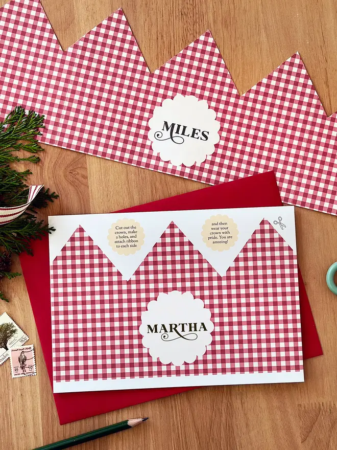 A personalised Christmas crown card with a red gingham design lays on a red envelope, on a wooden table.