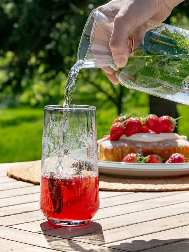 Water being poured into a tall, floral-etched highball glass filled with a red drink on an outdoor table.