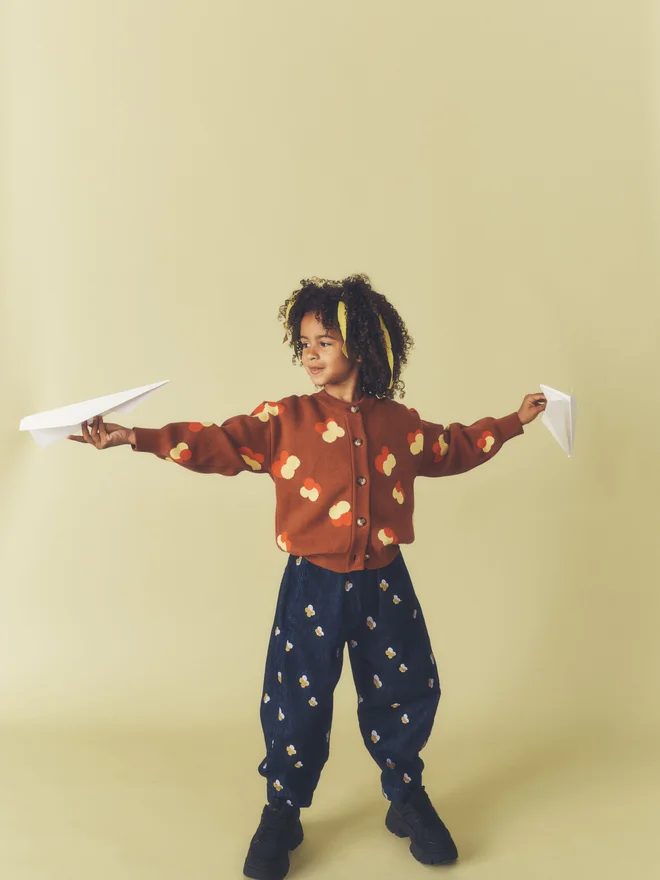 Child holds a paper plane in each outstretched hand against a pale yellow backdrop, wearing a Studio Koter chestnut floral intarsia cardigan and embroidered dark denim trousers.