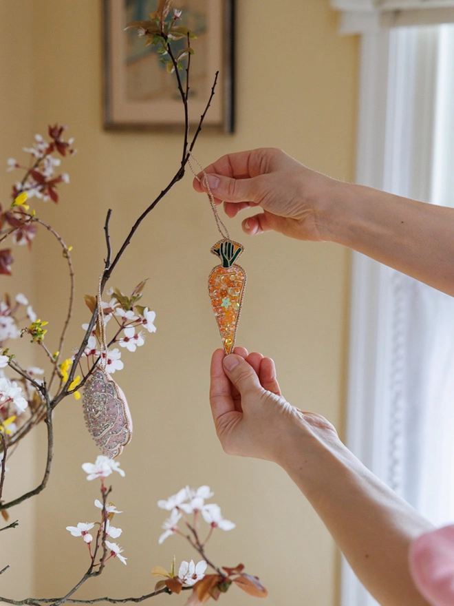 Embroidered sequin carrot decoration being hung by hand on a tree inside a house