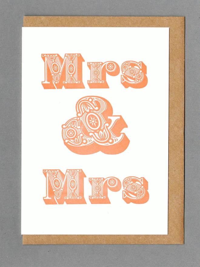 Orange text reading 'Mrs & Mrs' on white card with a brown envelope behind with a grey background