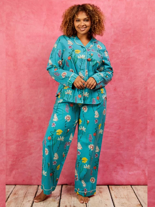 Model stands with her hands touching wearing a bright teal based pair of pyjamas, classic style with button down front and piping on shirt covered in purple, pink and yellow british wild flowers