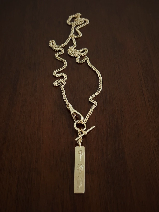 Gold plated sterling silver watch chain with a bar pendant to represent strength on a dark background