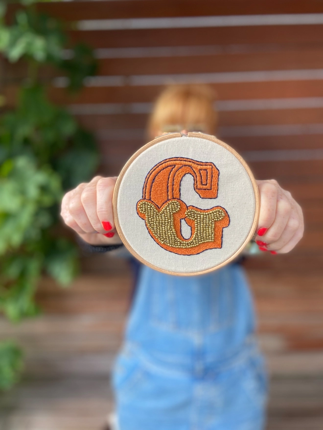 An embroidered hoop with a letter G, being held in front of a female 