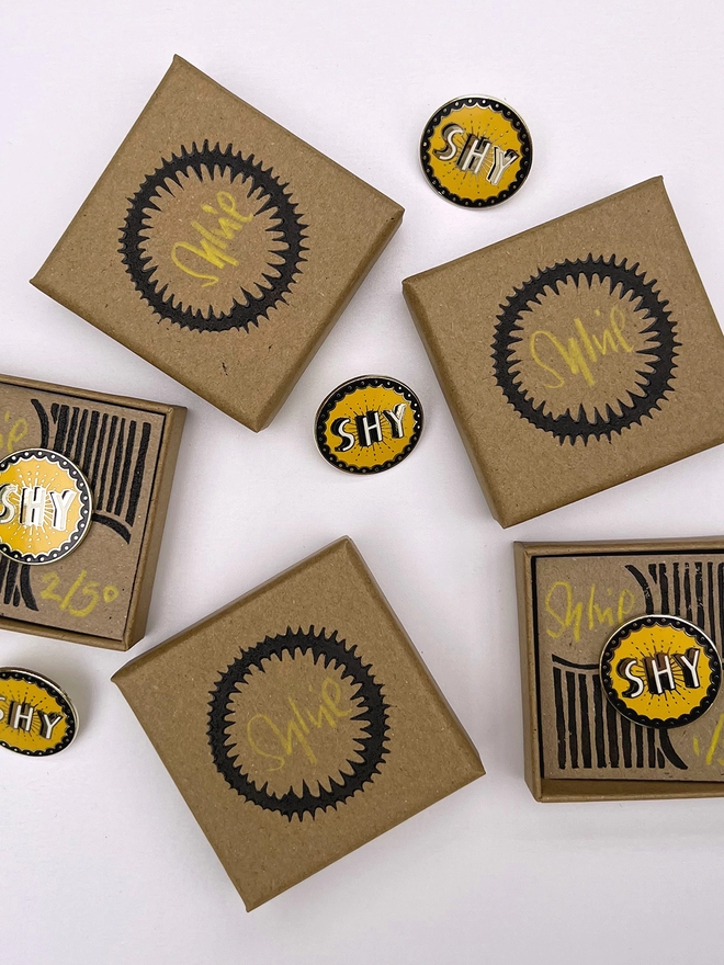 A close up of three SHY cloisonne brooches, they are mustard, black, white and brass coloured. Some of the brooches are in hand printed gift boxes.  