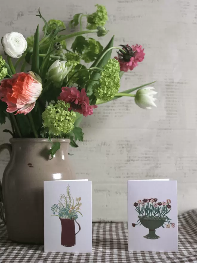 2 mini floral card next to vase of flowers on gingham tablecloth. 