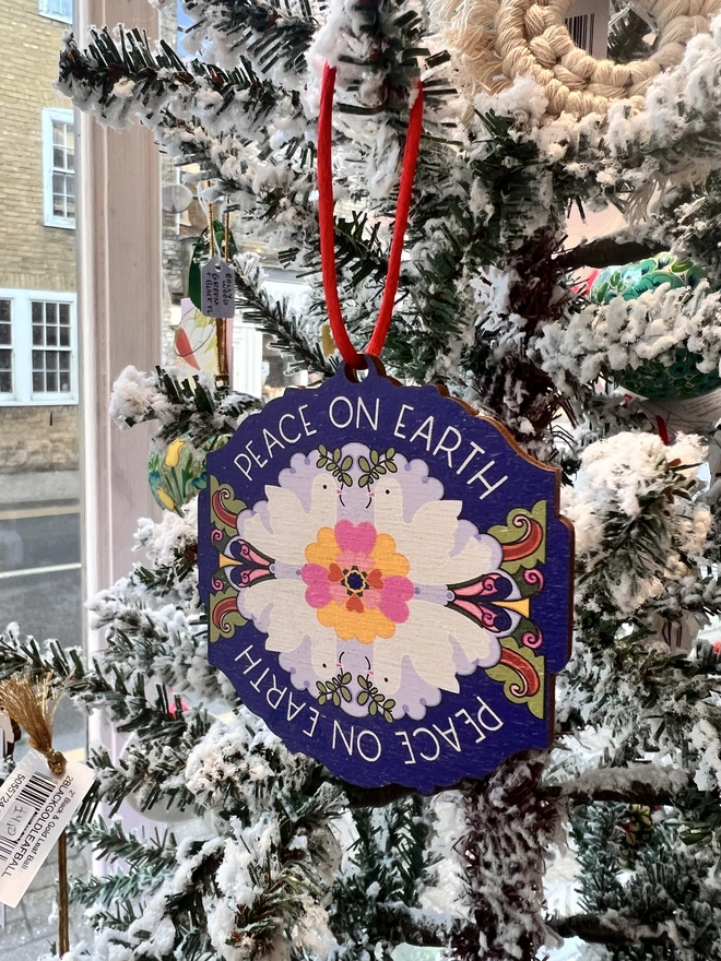 Multi coloured wooden tree decoration the reads 'Peace on Earth' around it, with four white doves and a flower pattern at the centre. It is hanging by a red ribbon on a fake white christmas tree with other decorations. 