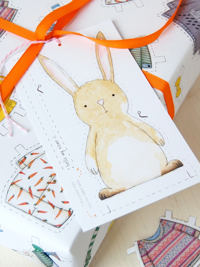 A gift wrapped in white wrapping paper with a hand illustrated outfit design and rabbit paper doll gift tags is beside a cut out rabbit paper doll on a white wooden table.