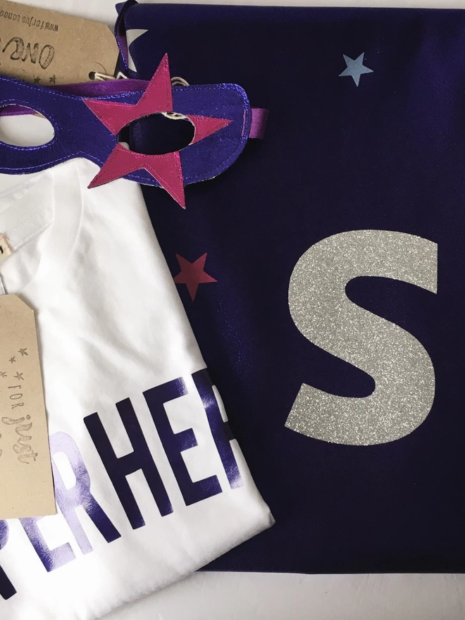 folded superhero costume, white T-shirt with superhero slogan in metallic purple, purple leather eye mask with pink star on the left eye, purple cape with silver glitter S initial