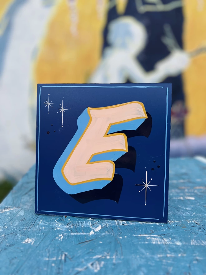 Hand-painted letter 'E' on a blue background with beiges and blues with a lined border.