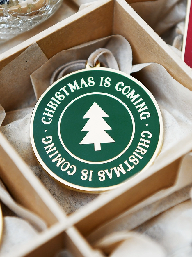 A deep green and gold enamel Christmas decoration, with the words Christmas Is Coming surrounding a gold Christmas tree, is tucked into a sectioned box.