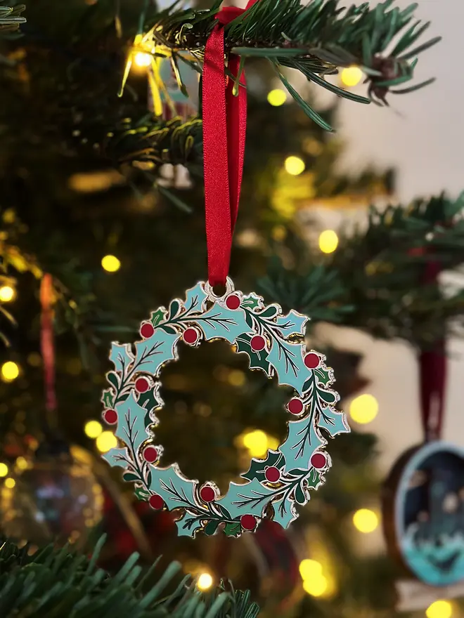 An heirloom holly wreath Christmas decoration hangs from a red ribbon on a decorated Christmas tree.
