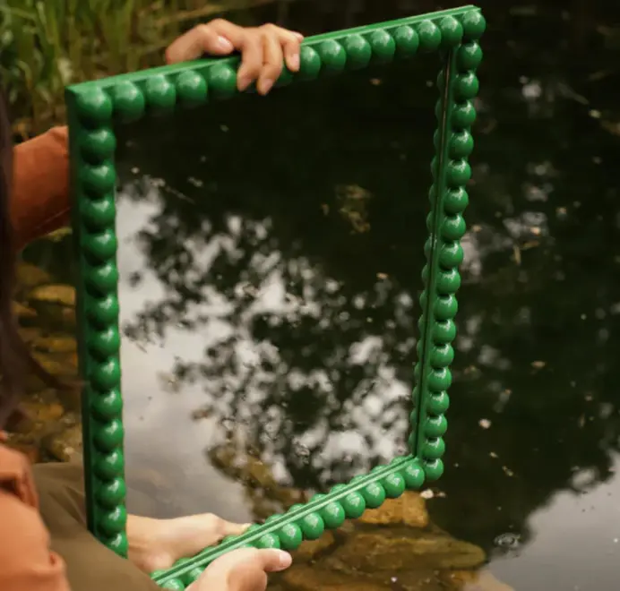 Over-sized bobbin mirror with a decorative turned-bead frame held in hands outdoors, reflecting trees in the mirror.