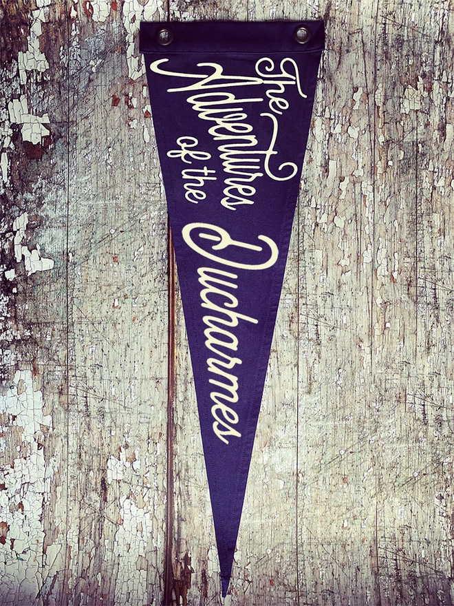 A navy coloured pennant flag hung vertically on a distressed wooden wall. In ivory lettering is the words ‘The Adventures of the Ducharmes'