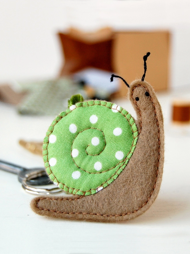 A homemade felt snail keyring with a green spotty cotton shell is on a white desk in front on the craft kit